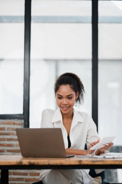 Bir kadın elinde dizüstü bilgisayar ve bir yığın kağıtla masada oturuyor. Gülümsüyor ve işinden zevk alıyor.