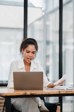 Modern bir ofiste dizüstü bilgisayarda çalışan genç bir kadın, elinde akıllı bir telefon, beyaz gömlek giyiyor, iş iletişimiyle meşgul..