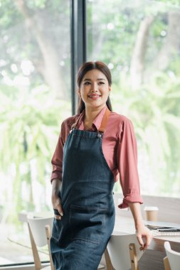 Kot önlüklü genç bir kadın modern bir kafede pencerenin önünde duruyor, gülümsüyor ve başka tarafa bakıyor..