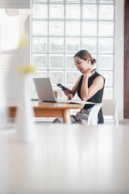 Bir kadın dizüstü bilgisayarı ve cep telefonu olan bir masada oturuyor. Cep telefonuna bakıyor ve bir şeye odaklanmış durumda.