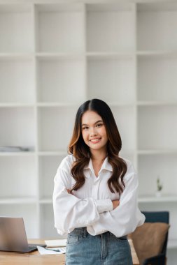 Gündelik giysiler içinde, modern ofis alanında kolları çapraz duran ve arka planda rafları olan kendinden emin genç bir kadın..