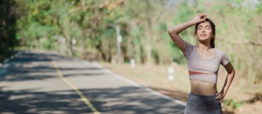 Bir kadın, eli alnında bir yolda yürüyor. Gri bir gömlek ve gri pantolon giyiyor.