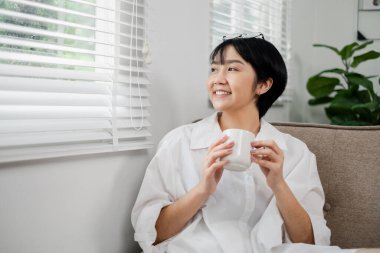 Parlak, modern bir oturma odasında, doğal ışık ve yeşil bitkilerle bir fincan kahvenin tadını çıkaran genç bir kadın..