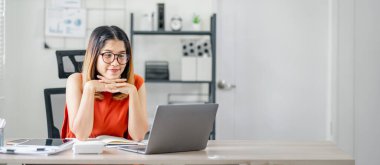 Önünde dizüstü bilgisayarı olan bir kadın masada oturuyor. O, gözlük takıyor ve elleri dizlerinin üzerinde. Sahne odaklanmış ve üretken bir atmosferi gösteriyor.