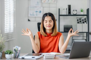 Turuncu gömlekli bir kadın elinde dizüstü bilgisayar ve kitapla bir masada oturuyor. Yoga yapıyor ve sakin ve odaklanmış durumda.