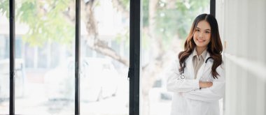 Beyaz önlüklü bir kadın kollarını kavuşturmuş bir pencerenin önünde duruyor. Yüzünde bir gülümseme var ve olay yerinde bir doktorun resmi var.