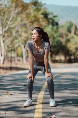 Spor yapan genç bir kadın güneşli bir günde arka planda ağaçlar ve dağlarla güneşli bir parkta spor yaparken dinleniyor..