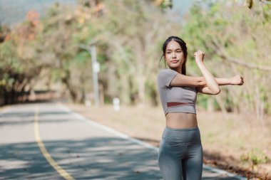 Spor yapan genç bir kadın güneşli bir günde kollarını açık havada ağaçlar ve arka planda patika olan bir parkta esnetiyor..