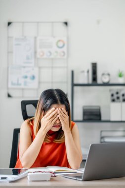 İş kadını masasında, elleri yüzü üzerinde, modern bir ofis ortamında dizüstü bilgisayar ve belgelerle stresli hissediyor. İşyeri stresi ve kurumsal baskı kavramı.