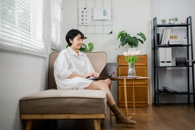 Oturma odasındaki bir kanepede oturan bir kadın dizüstü bilgisayarıyla çalışıyor. Gülümsüyor ve işinden zevk alıyor. Oda saksı bitkileri ve bir vazo çiçekle süslenmiş, rahat bir ortam sağlıyor.