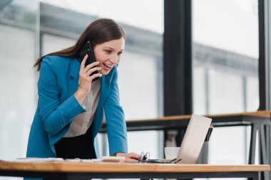 Bir kadın masada otururken cep telefonuyla konuşuyor. Mavi bir ceket ve beyaz bir gömlek giyiyor.