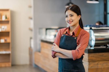 Mavi önlüklü bir kadın bir restoranda yüzünde bir gülümsemeyle duruyor. Kırmızı bir gömlek giyiyor ve kollarını kavuşturuyor.