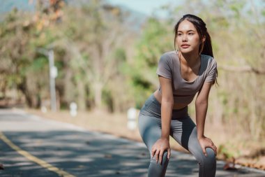 Gri gömlekli ve gri pantolonlu bir kadın yolda duruyor. Kameraya ciddi bir ifadeyle bakıyor.