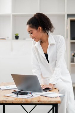 Modern bir ofiste dizüstü bilgisayarla çalışan, beyaz kıyafetli, genç, profesyonel bir kadın. Minimalist dekorasyonu ve doğal ışığı var..