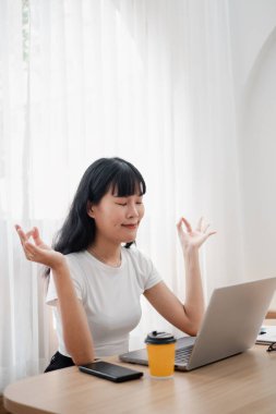 Genç bir kadın, elinde bir dizüstü bilgisayar ve kahve bardağıyla masa başında oturmuş modern bir ofis ortamında meditasyon yapıyor ve farkındalık yapıyor..