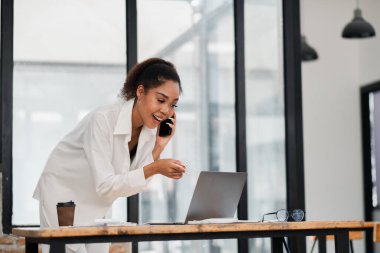 Modern bir ofiste iş iletişimi için dizüstü bilgisayar ve akıllı telefon kullanan profesyonel bir kadın..