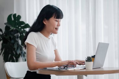 Modern bir ev ofisinin dizüstü bilgisayarında çalışan genç bir kadın doğal ışık ve kapalı bitkilerle, günlük beyaz bir tişört giyiyor, üretken ve rahat bir uzaktan çalışma ortamını temsil ediyor..