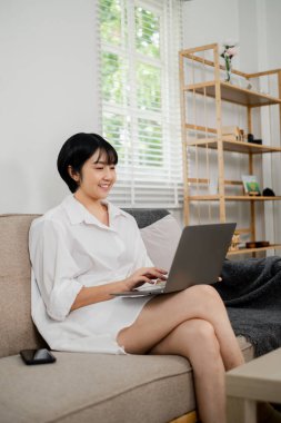 Parlak, modern bir oturma odasında dizüstü bilgisayar kullanan gülümseyen genç bir kadın doğal ışık ve şık dekorasyonlu bir odada oturuyor..