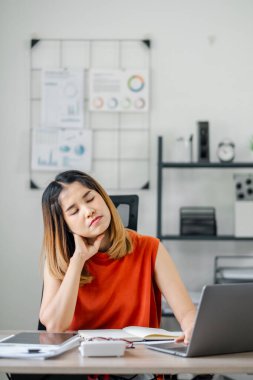 Masasında dizüstü bilgisayarı olan kırmızı elbiseli bir kadın, modern ofis ortamında boyun ağrısı ve yorgunluk yaşıyor..