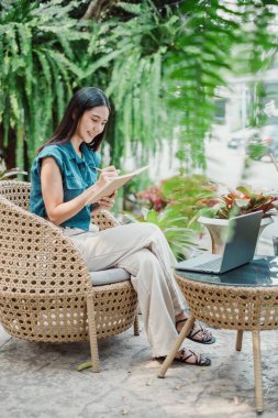 Açık hava bahçesinde tablet kullanan genç bir kadın. Hasır mobilyaların üzerinde oturuyor. Laptopuyla..
