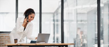 Modern bir ofiste çalışan profesyonel bir kadın, bir telefonla birden fazla iş yapıyor ve bir dizüstü bilgisayarla çalışıyor, üretken bir çalışma ortamı sergiliyor..