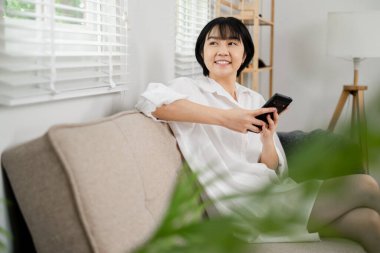 Beyaz gömlekli bir kadın, elinde akıllı bir telefonla bej bir kanepede oturuyor, gülümsüyor ve modern bir oturma odasına bakıyor..