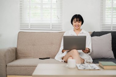 Genç bir kadın parlak bir oturma odasında koltukta otururken dizüstü bilgisayar kullanıyor. Kahve ve kitaplarla dolu rahat ve rahat bir ev..