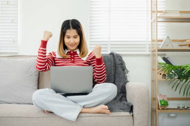 Gündelik kıyafetlerle başarıyı kutlayan genç bir kadın evde dizüstü bilgisayarla çalışırken, parlak ve modern bir oturma odasında bir kanepede otururken..