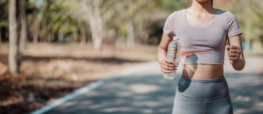 Parkta koşan, elinde su şişesiyle koşan, atletik kıyafetler giyen, güneşli bir günde dışarıda egzersiz yapan genç bir kadın..