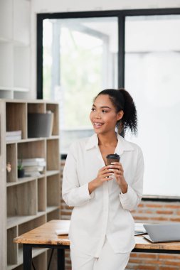 Modern ofiste elinde kahve fincanı tutan, gülümseyen ve yüzünü çeviren, beyaz iş kıyafeti giymiş genç profesyonel bir kadın..