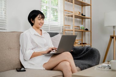 Genç bir kadın oturma odasındaki koltukta dizüstü bilgisayar kullanıyor. Rastgele ve rahat ev ortamları, uzak çalışma ya da öğrenim için ideal.