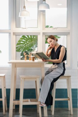 Genç bir kadın modern minimalist bir kafede yüksek bir masada oturuyor ve arka planda doğal ışık ve yeşillik olan bir kitap okuyor..
