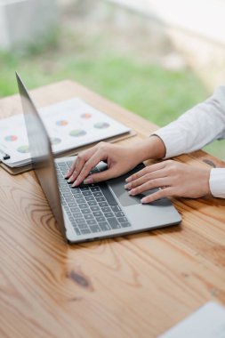 Parlak bir ofis ortamında, ahşap bir masada, üzerinde iş belgeleri olan bir dizüstü bilgisayarın klavyesinde yazı yazan eller..