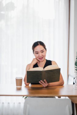 Ahşap bir masada kitap okuyan bir kadın. Parlak, modern bir odada, perdeli bir kahve fincanıyla..