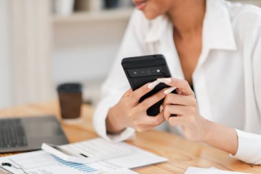 Modern bir ofiste belgelerle ve kahveyle bir masada çalışırken elinde akıllı telefon olan bir iş kadınının yakın çekimi..