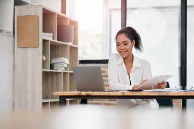 Modern bir ofiste dizüstü bilgisayarda çalışan gülümseyen genç bir kadın. Elinde bir dosya var ve odaklanmış ve meşgul görünüyor..