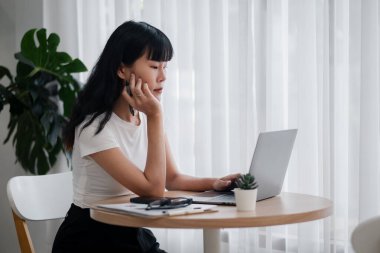 Beyaz gömlekli genç bir kadın yuvarlak ahşap bir masada dizüstü bilgisayarda çalışıyor, doğal ışık perdelerin arasından akıyor ve arka planda iç mekan bitkileri var..
