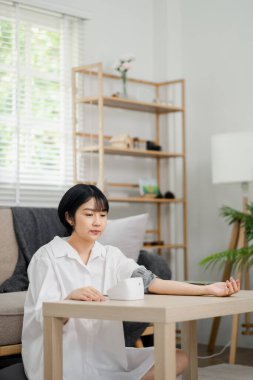 Beyaz gömlekli genç bir kadın, modern dekorlu minimalist bir oturma odasında otururken kan basıncı monitörü kullanıyor..