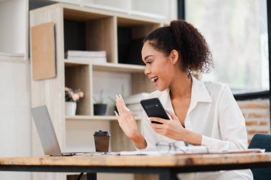 Sanal görüşmede genç bir kadın elinde akıllı telefon tutarken dizüstü bilgisayara gülümsüyor ve el sallıyor. Doğal ışık ile modern çalışma alanı.