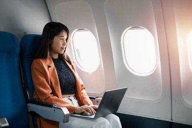 Uçuş sırasında dizüstü bilgisayarda çalışan iş kadını, bir uçak kabininde oturmuş, pencerelerden güneş ışığı geçiyor..