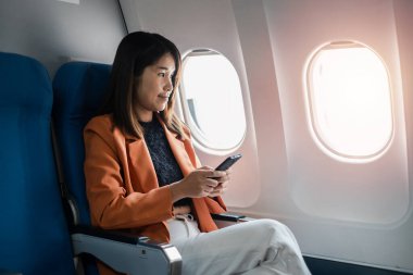 Pencere kenarında oturan turuncu ceketli genç bir kadın uçakta akıllı telefon kullanıyor. Modern hava yolculuğu konsepti.