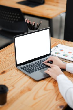 Modern ofis ortamında boş bir ekranla dizüstü bilgisayarda yazan eller, tahta bir masa, grafikli bir pano ve kahve fincanı..