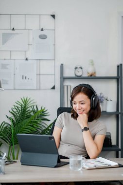 Kulaklık takan genç bir kadın, tablet üzerinde çalışıyor ve arka planda bitki ve raflarla modern bir ev ofisinde gülümsüyor..