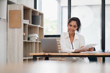 Modern ofis ortamında genç profesyonel bir kadın, bir masada dizüstü bilgisayar ve not panosuyla çalışıyor, güvenle gülümsüyor..