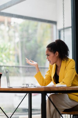 Sarı ceketli profesyonel bir kadın büyük pencereli çağdaş bir ofis ortamında dizüstü bilgisayarında hayal kırıklığını gösteriyor..
