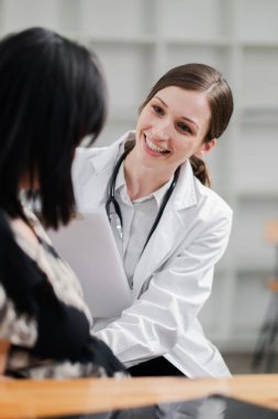 Beyaz önlüklü, gülümseyen kadın doktor bir hastaya danışmanlık yapıyor, modern bir muayenehanede elinde bir pano tutuyor..