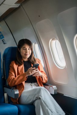 İş kadını uçakta seyahat ederken akıllı telefon kullanıyor, pencere kenarında oturuyor, turuncu ceket ve beyaz pantolon giyiyor..