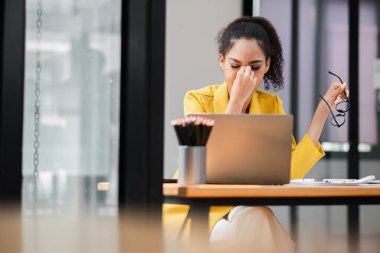 Sarı ceketli genç bir iş kadını modern bir ofiste dizüstü bilgisayarıyla çalışıyor. Stresli ve yorgun hissediyor..