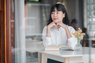 Genç bir kadın rahat bir kafede bir masada oturup kitap okuyor. Kafede minimalist dekor ve doğal ışık yer alıyor..