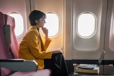 Sarı ceketli profesyonel bir kadın uçuş sırasında dizüstü bilgisayarda çalışıyor, uçak penceresinin yanında oturuyor, ekrana odaklanıyor..